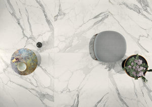 Three decorative items on a marble look tile surface: a round metallic object, a gray textured sphere, and a circular plant pot with flowers.