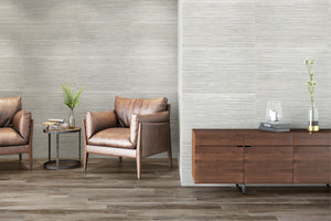 A modern interior scene showing textured light gray walls, medium-brown wood flooring, a brown leather armchair, a matching side table with a plant, and a dark wood credenza with decorative objects.