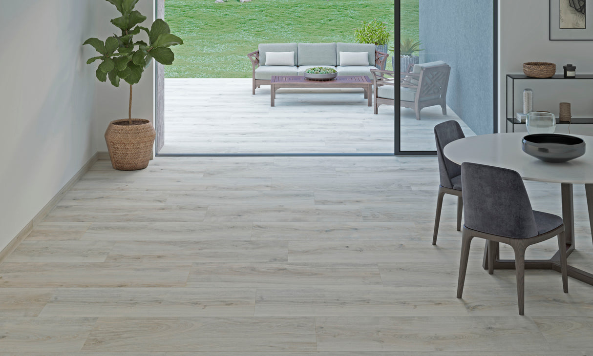 Light-colored wood-look tile flooring installed in a modern interior room, with a dining table and chairs on the right and an open doorway leading to a patio and green lawn.
