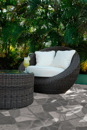 A dark woven round lounge chair with white cushions sits on a stone patio, accompanied by a matching round side table holding a drink, with dense tropical green foliage in the background.
