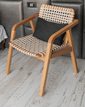 A wooden armchair with woven beige and white rope seat and back, styled with a dark gray throw pillow, set against a gray upholstered headboard and light wood floor.