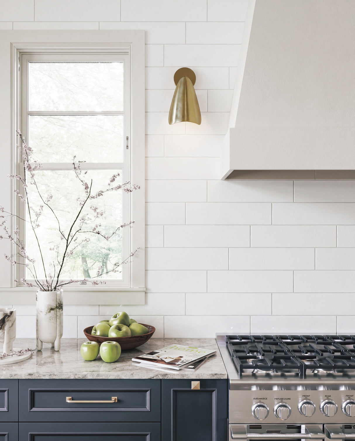 A modern kitchen scene with white subway tile walls, a brass wall sconce, a gas range, and a marble countertop holding green apples and a vase with branches.