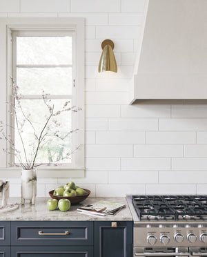 A modern kitchen scene with white subway tile walls, a brass wall sconce, a gas range, and a marble countertop holding green apples and a vase with branches.