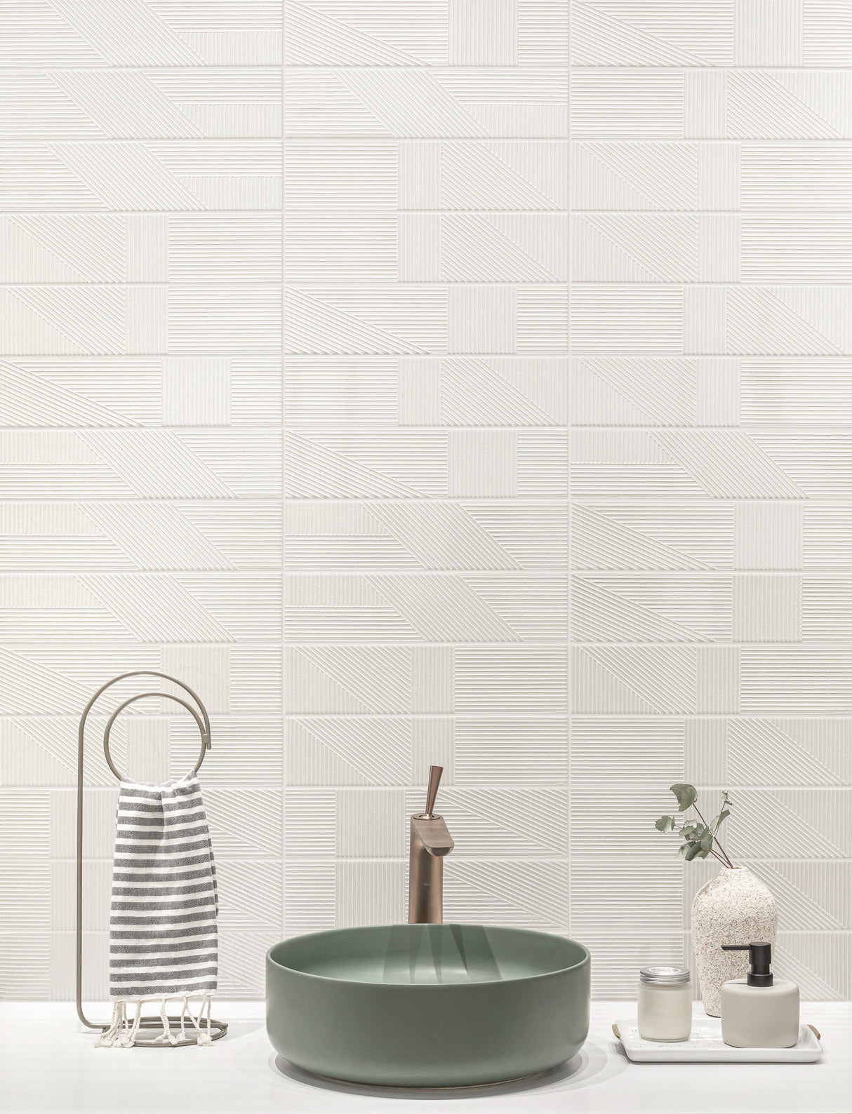 A styled bathroom scene featuring a green vessel sink with a bronze faucet, set against a white textured wall with horizontal tiles. To the left is a striped towel on a metal ring holder, and to the right are small decorative bottles and a vase.