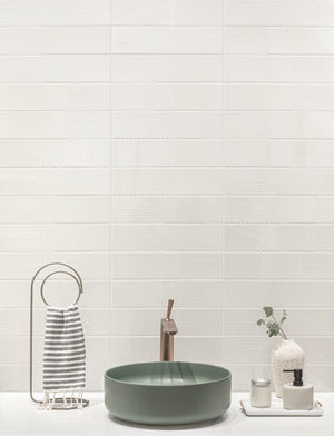 A styled bathroom scene featuring a green vessel sink with a bronze faucet, set against a white textured wall with horizontal tiles. To the left is a striped towel on a metal ring holder, and to the right are small decorative bottles and a vase.