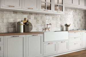 A modern kitchen interior with white shaker-style cabinets, a glossy light gray tile backsplash, wood countertops, and a white farmhouse sink with a copper faucet.