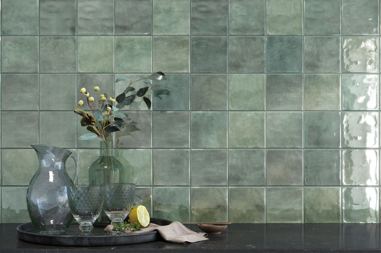 Green square ceramic wall tiles with a glossy, textured finish, styled in a kitchen setting with a dark countertop, glassware, and a vase of eucalyptus.