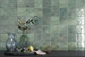 Green square ceramic wall tiles with a glossy, textured finish, styled in a kitchen setting with a dark countertop, glassware, and a vase of eucalyptus.