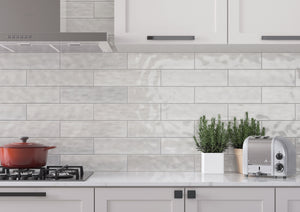 Light gray subway tile backsplash installed on a kitchen wall, shown with white cabinets, a gas stove with a red pot, potted green plants, and a silver toaster.