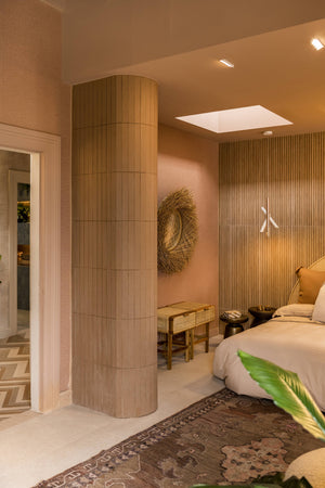 A modern bedroom interior with warm beige and blush tones, featuring a tall cylindrical column, a slatted wood accent wall, a bed with white bedding, and a patterned rug.