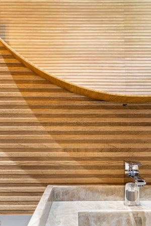 A close-up of a modern bathroom sink with a chrome faucet set into a light stone basin, set against a curved wooden slat wall.