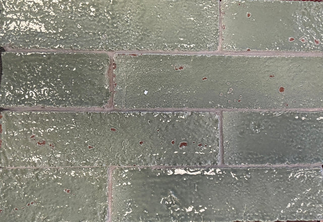Close-up of Artesano Tulum Sage 2x9 glossy clay brick tile in running bond, rippled sage-green glaze with red clay chips